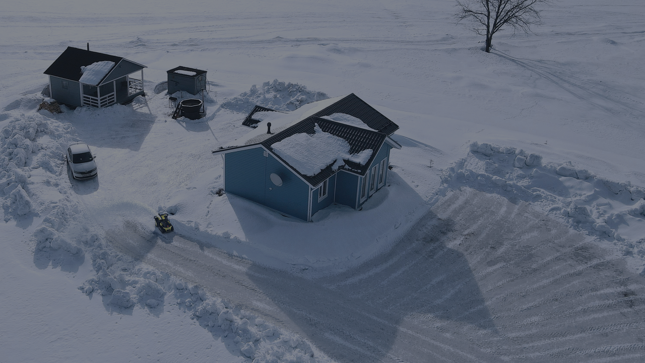 Aerial view of a snow-covered blue house with cleared driveway, snowbanks, and a small snow blower, showcasing winter maintenance for homeowners.