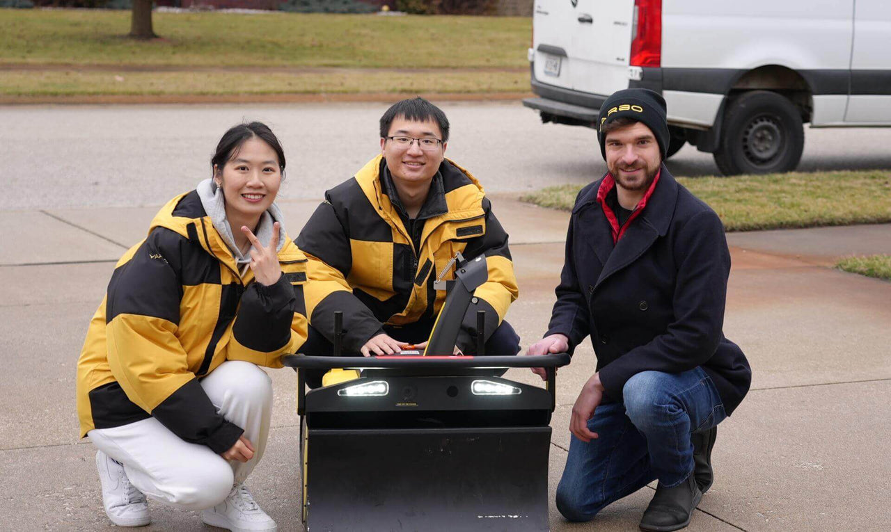 Three people in winter gear crouch beside a robotic device, smiling warmly outdoors near a white van.
