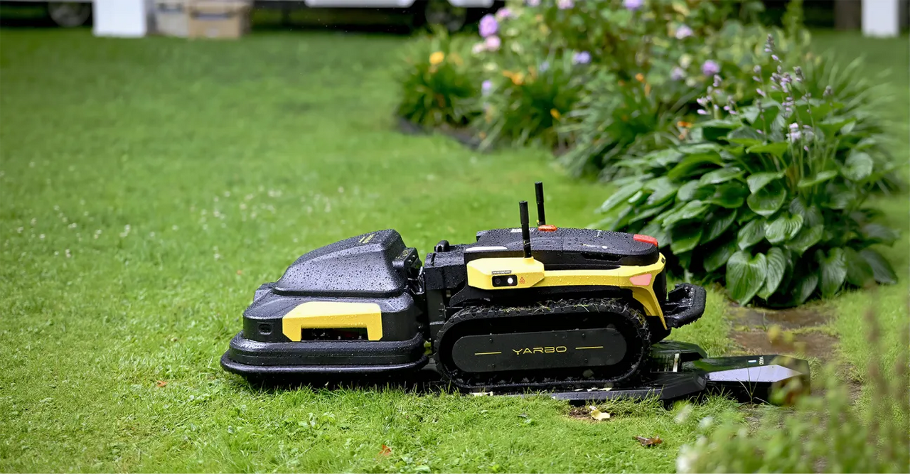 Black and yellow robotic lawn mower on wet grass near flowerbed, paused mid-task in a lush garden.