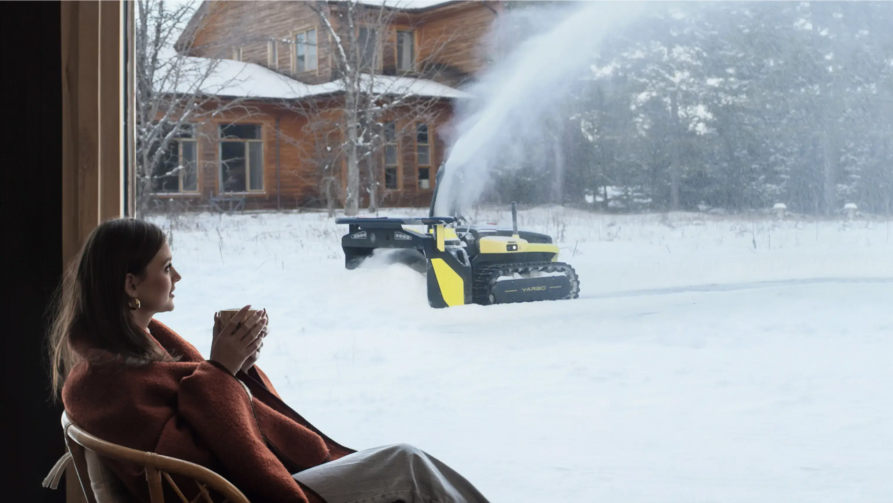 Woman sipping tea indoors watches robotic snow blower clear driveway outside snowy cabin.