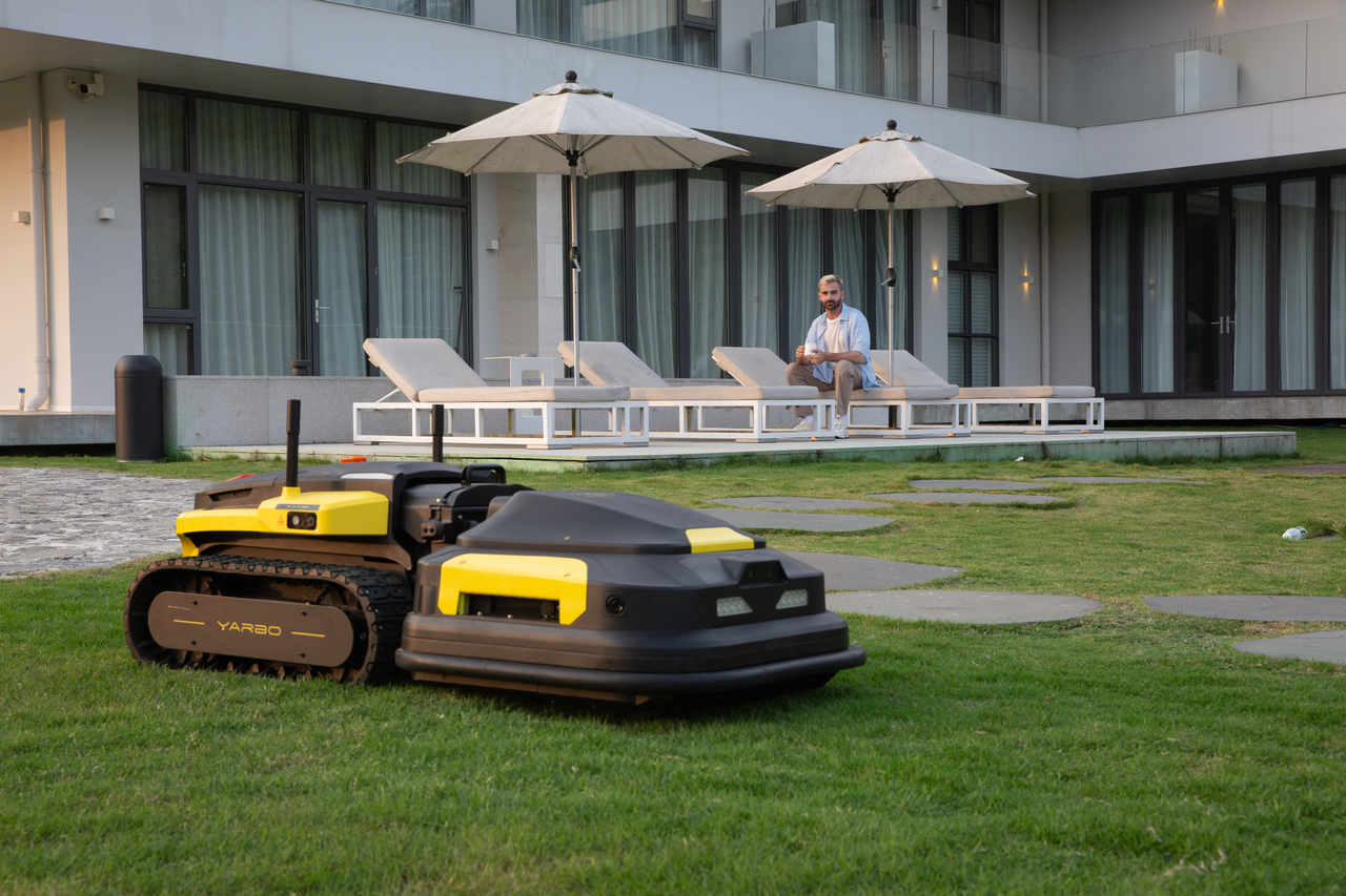 Two robotic lawn mowers on a lush green lawn, with a man relaxing on a lounge chair under umbrellas near a modern home.