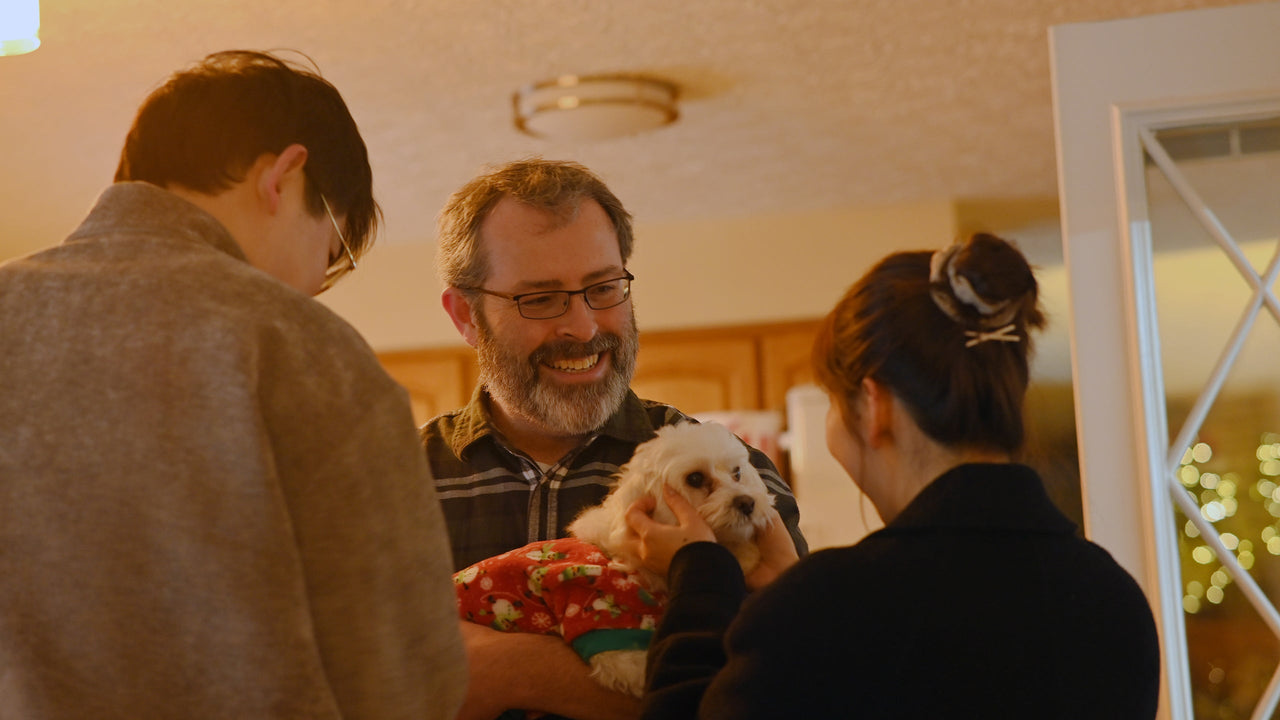 Smiling man holds fluffy white dog in festive sweater, surrounded by family in warm, cozy home.