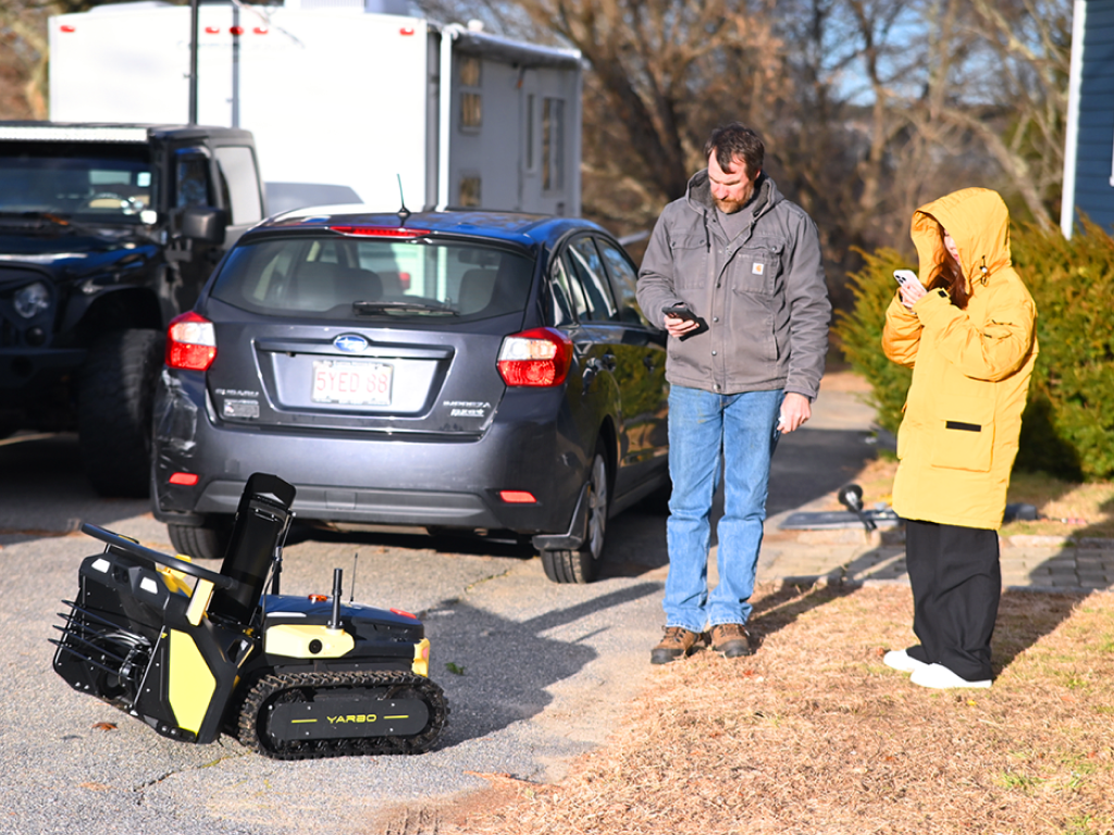John Burns tests Yarbo robot outdoors with assistant, checking device near parked cars on sunny day.