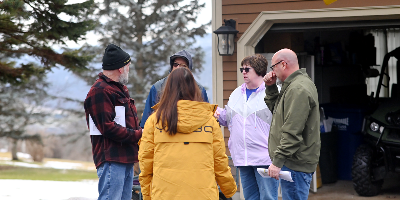 Kent Ross and guests gather outdoors in winter gear, discussing plans near a garage with snowy trees in the background.