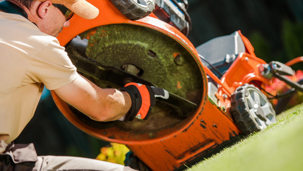 Man in gloves cleans rusted orange lawn mower on grass, preparing for safe disposal or recycling.