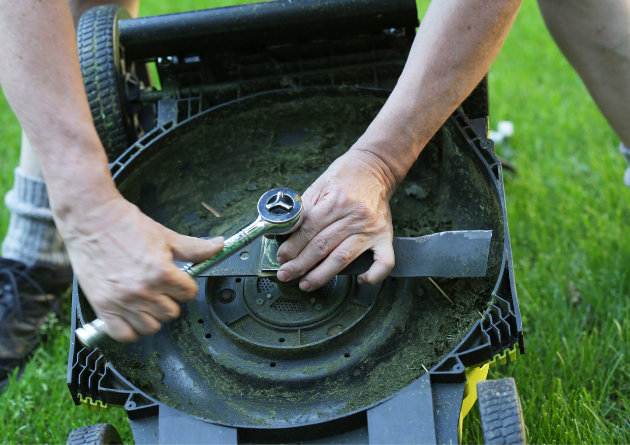 Hands using a wrench to remove a lawn mower blade from a grass-covered deck, showing maintenance in action.