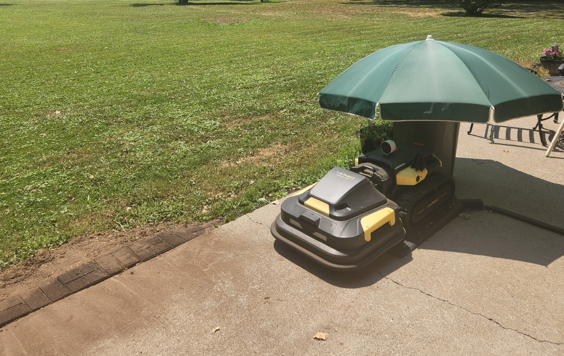 Robotic lawn mower under green patio umbrella on concrete patio, protected from sun and weather.