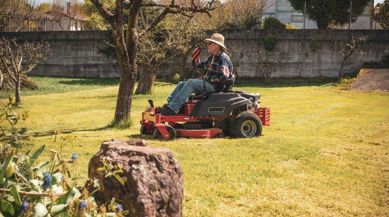 Man in straw hat sipping drink while riding red lawn mower across sunny, tree-dotted yard.