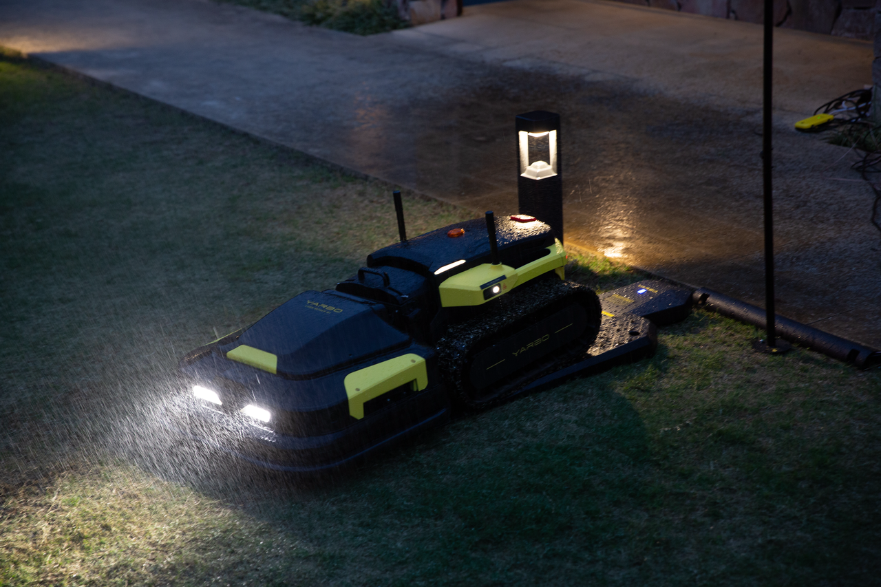 Robot lawn mower with glowing headlights operates on wet grass at night, charging station nearby.