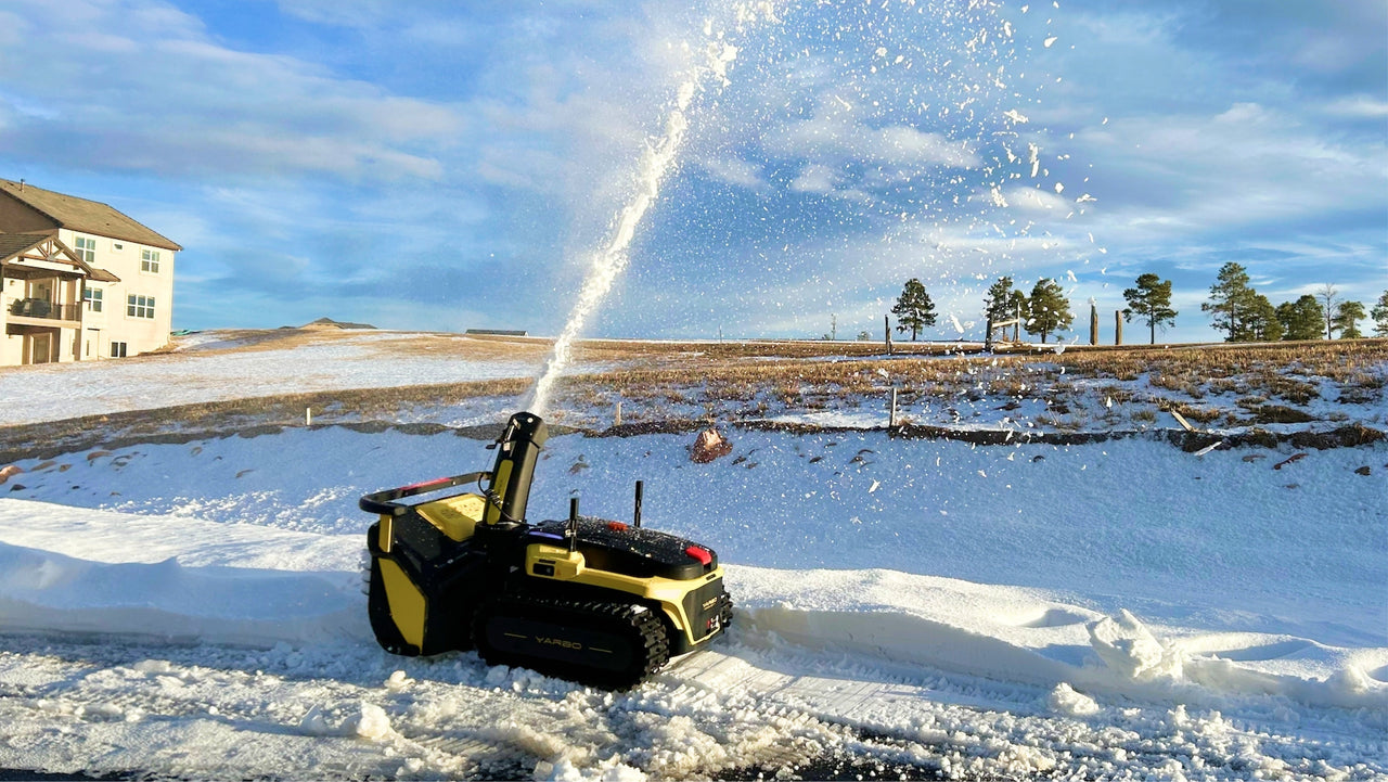 Yellow robotic snow blower clears heavy wet snow in a bright winter yard, spraying snow into the air.