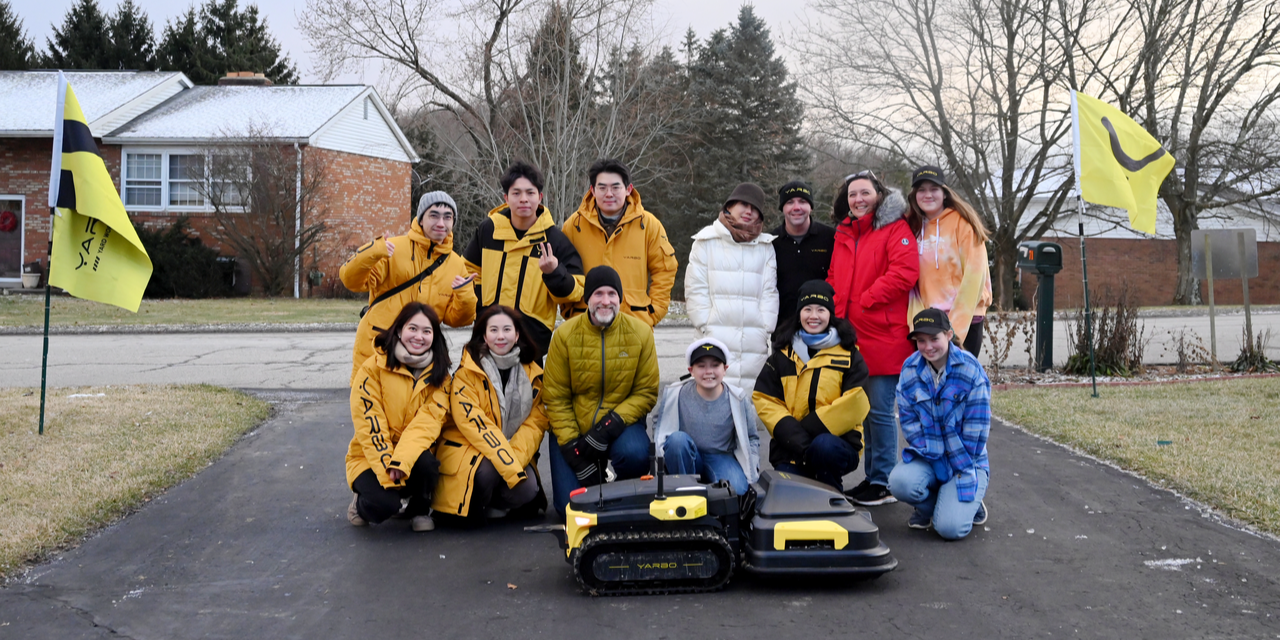 Group in yellow Yarbo jackets poses with robotic devices on a driveway, flanked by branded flags, showcasing a tech-driven team experience.