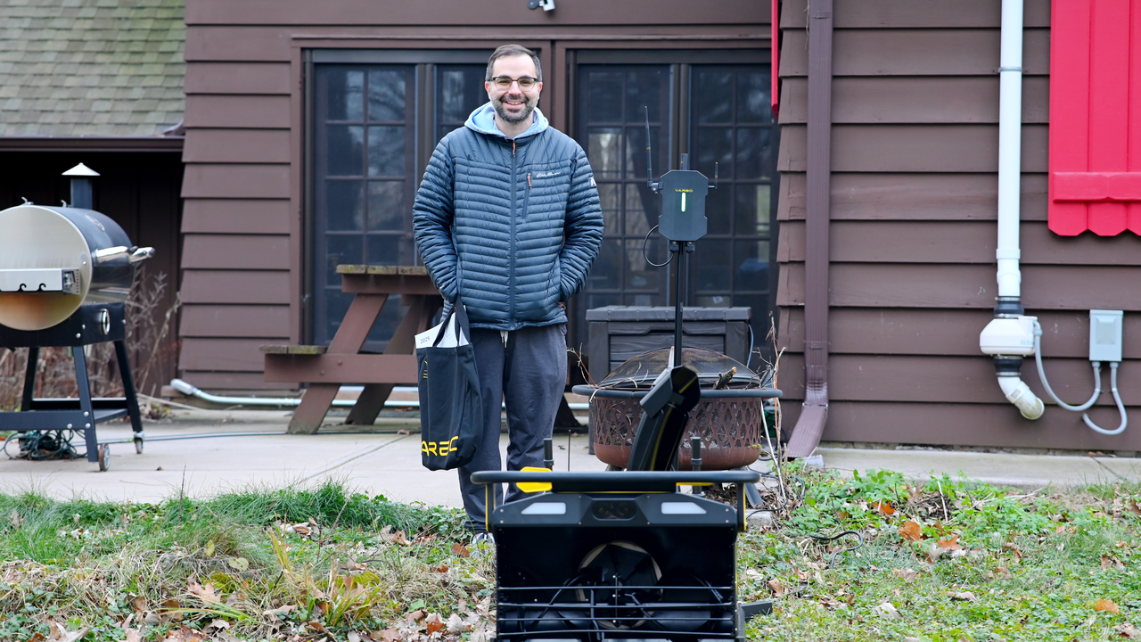 Man in puffer jacket stands proudly beside Yarbo robotic lawn mower in backyard, smiling with smart yard care gear.