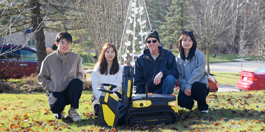 Four people pose with a yellow and black Yarbo robot lawn mower in a sunny Indiana yard, smiling amid fallen leaves.