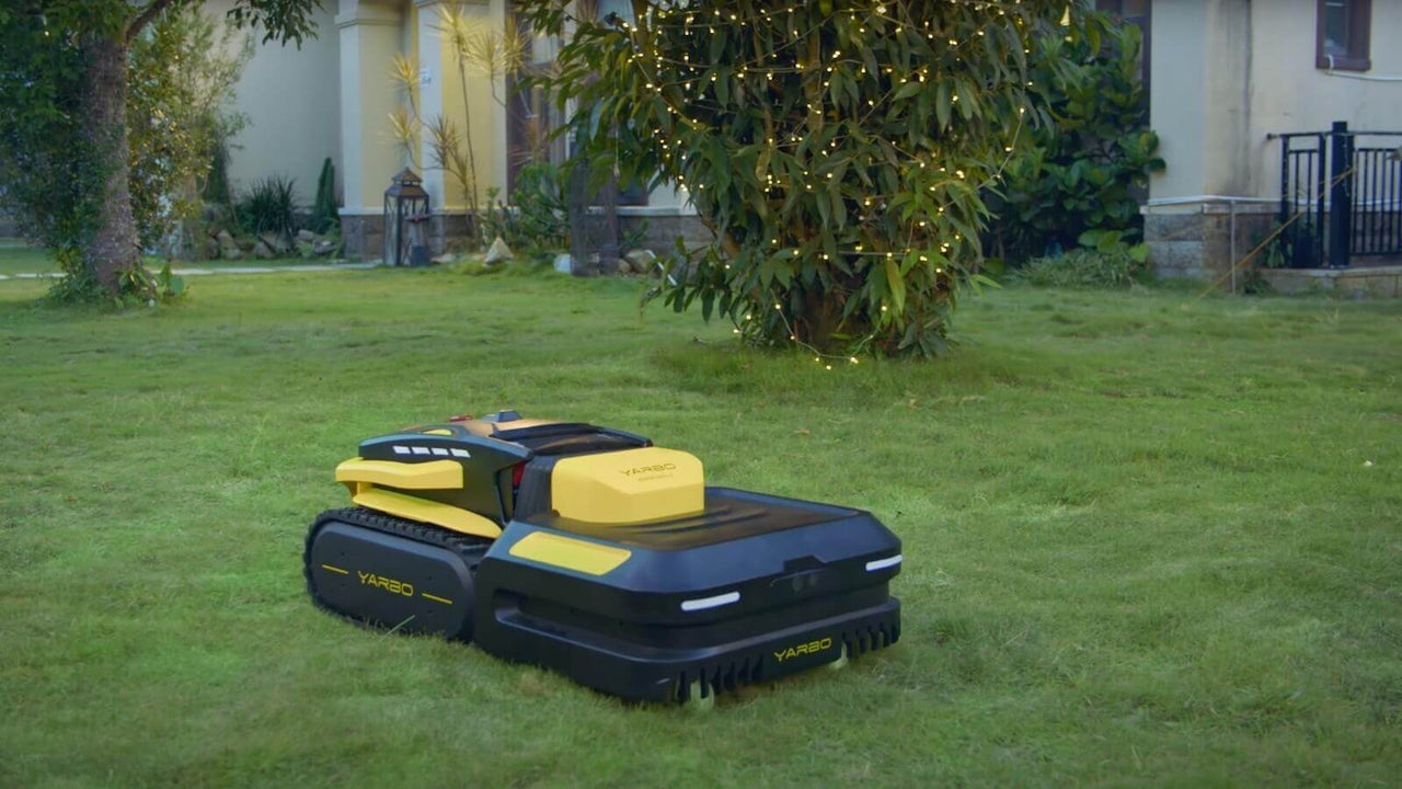 Black and yellow YARBO robot lawn mower on lush green grass near a tree with fairy lights, evening setting.