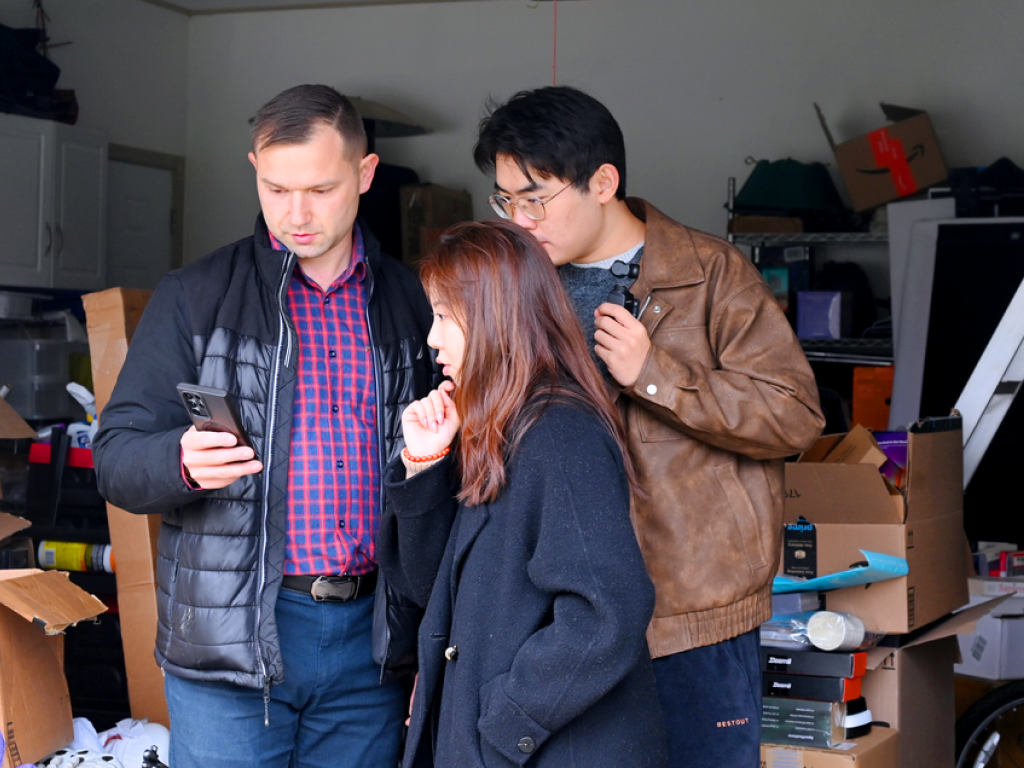 Three people huddle in a cluttered garage, focused on a phone screen, sharing a moment of collaboration and curiosity.