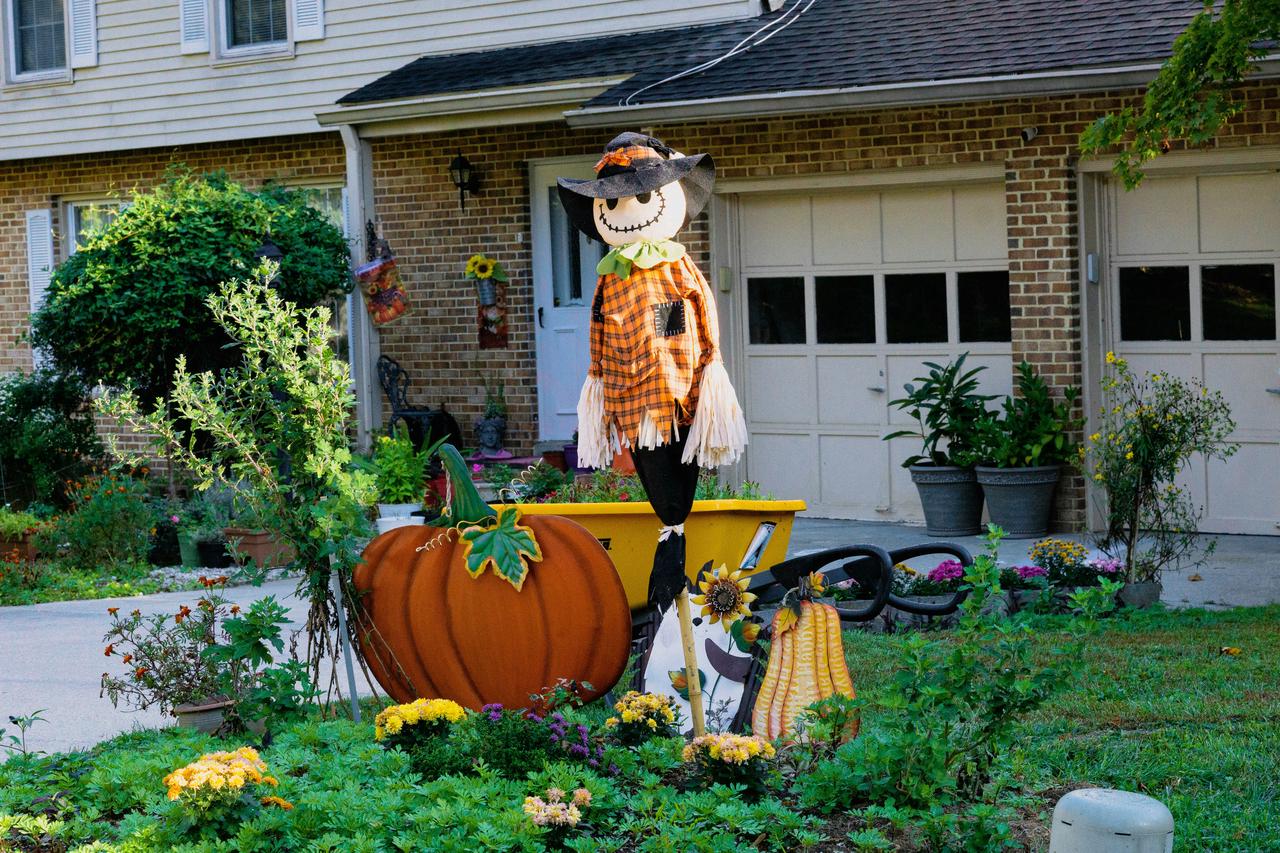 Cheerful Halloween yard display with scarecrow, pumpkin, and flowers in front of a cozy brick home.