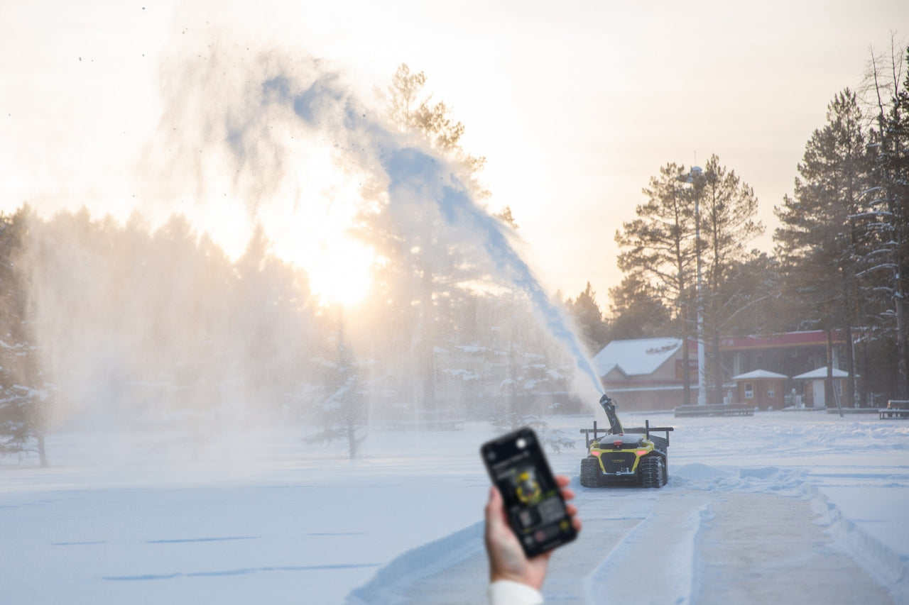 Hand holds phone controlling Yarbo robot spraying snow in winter, sunlit forest backdrop, showcasing reliable remote operation.