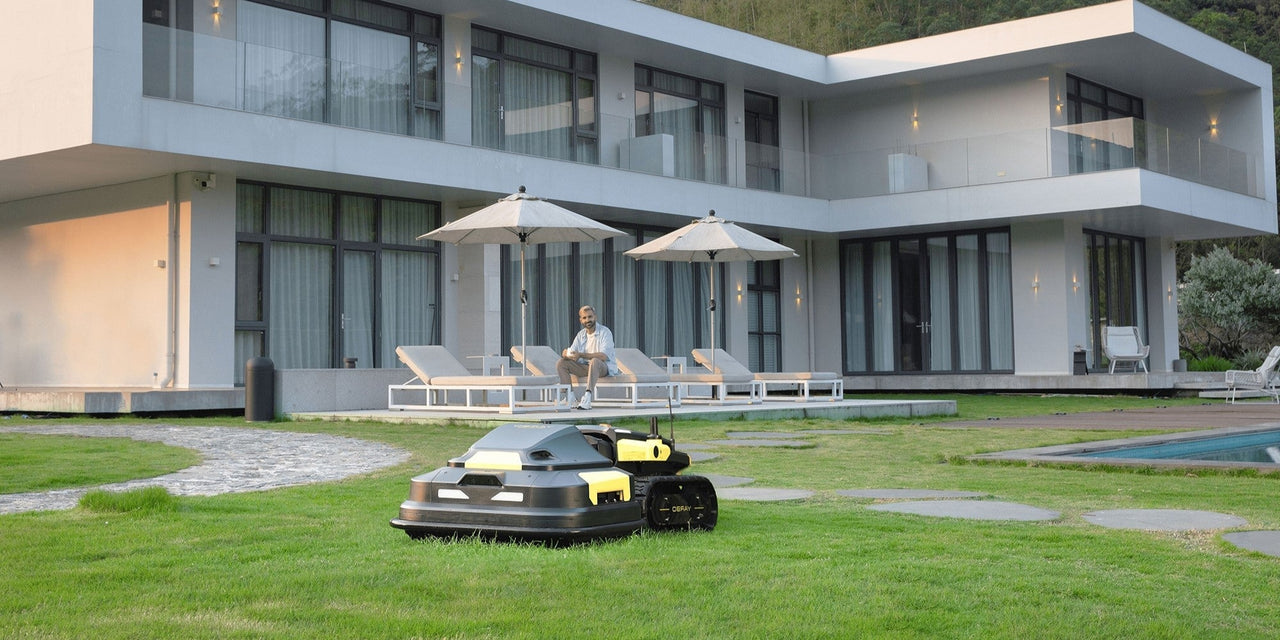 Robot lawn mower on vibrant green grass near modern home, man relaxing nearby under umbrellas.