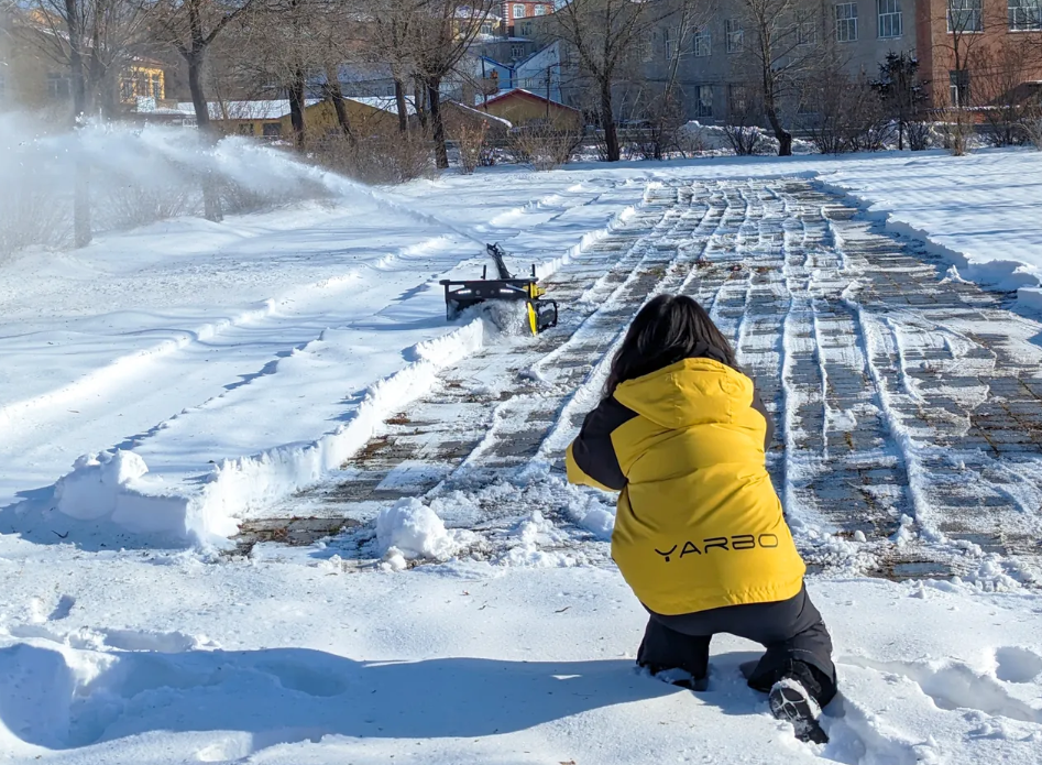 Person in yellow Yarbo jacket kneels beside snow-clearing machine, efficiently removing ice from a snowy brick path.
