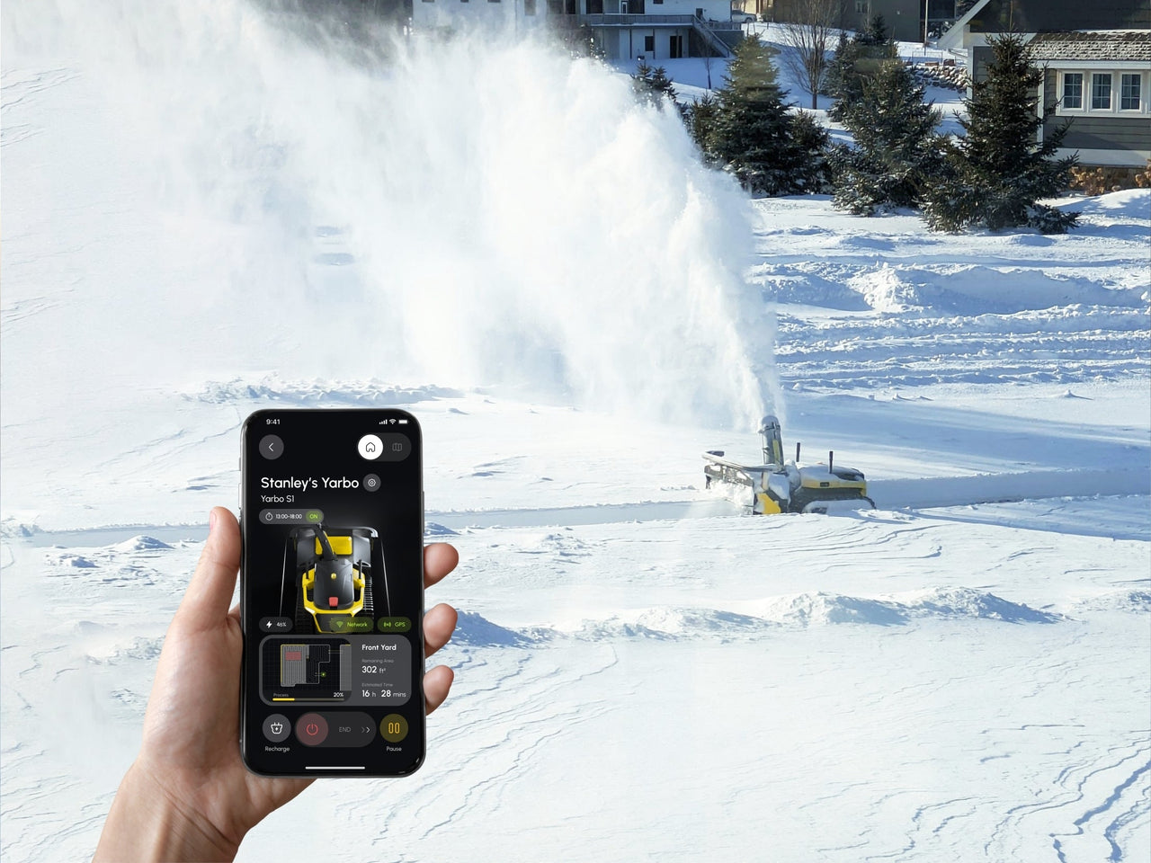Hand holds phone controlling robotic snow blower clearing snowy yard, showing app interface and snow spray.
