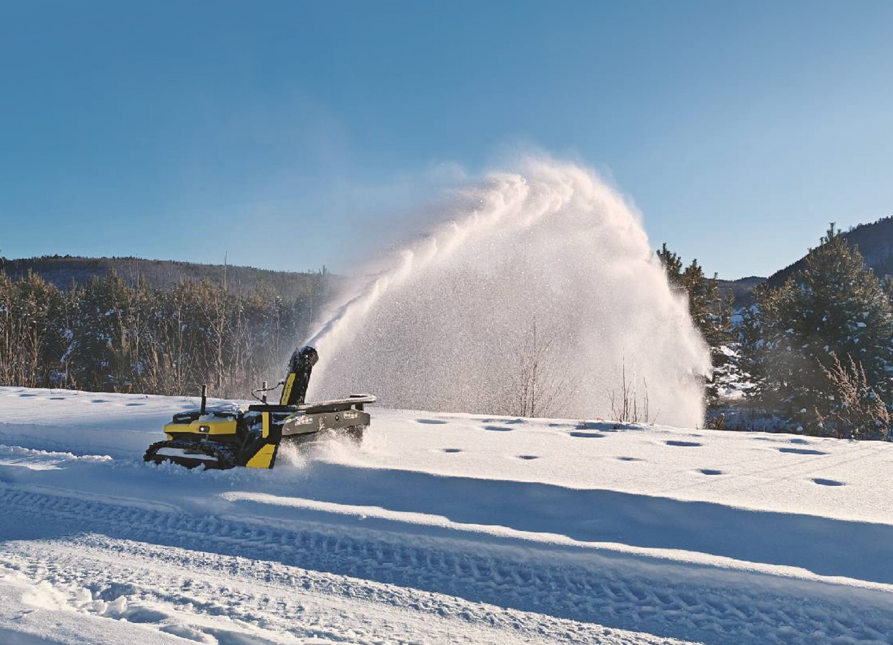 Yellow snow blower clearing path in snowy field, spraying snow into air under bright blue sky.