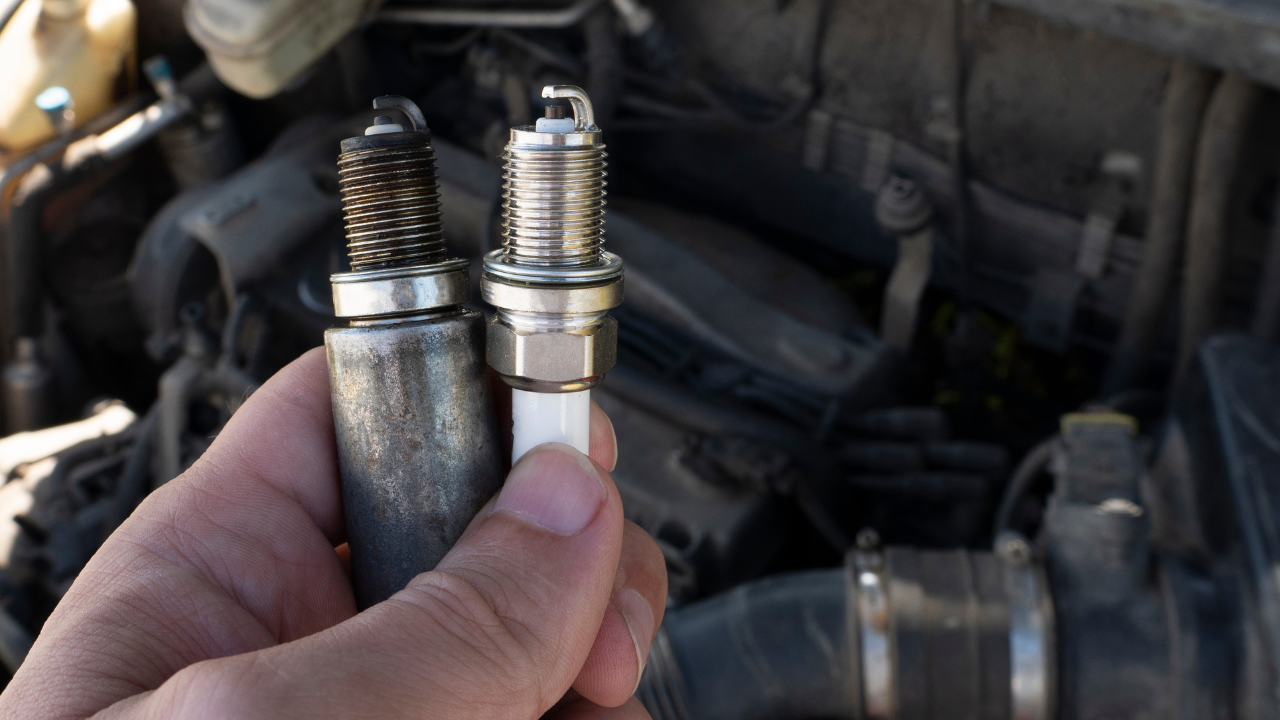 Hand holds worn and new spark plugs side by side, lawn mower engine in background, highlighting replacement need.