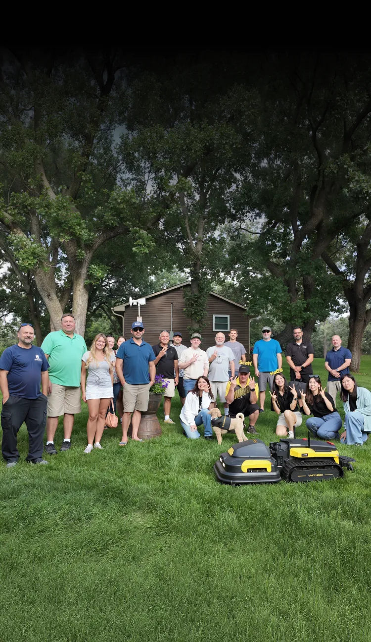 Group of people posing outdoors on grass with two yellow and black robotic mowers in foreground, trees and shed in background.