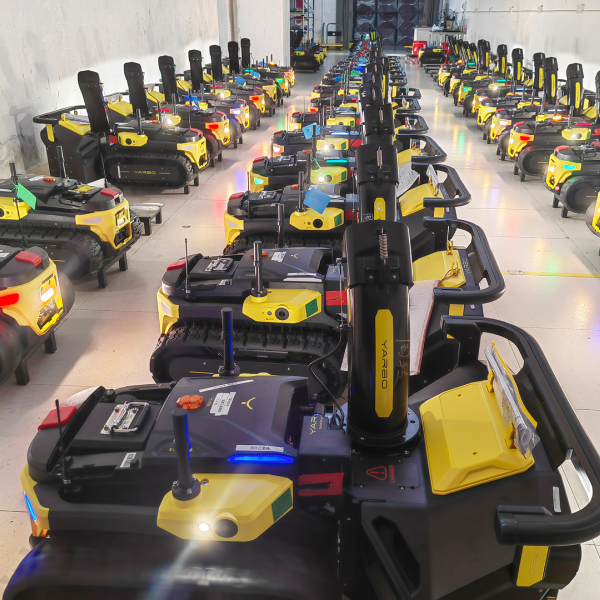 Rows of yellow and black Yaaro robotic lawn mowers lined up in a warehouse, illuminated by bright overhead lights.