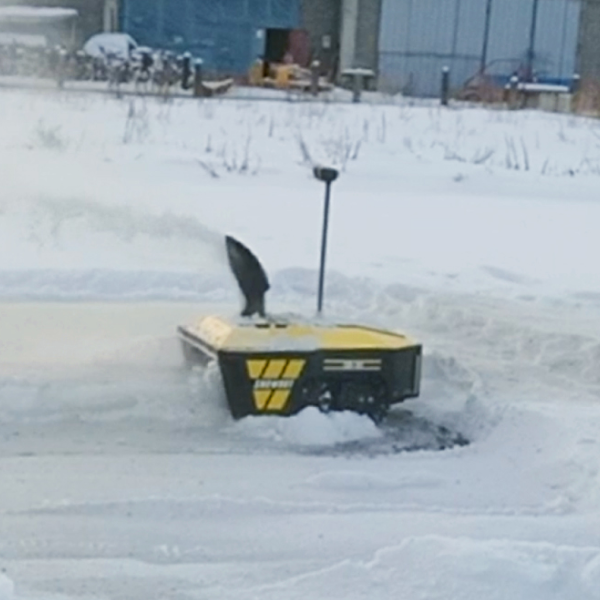 Yellow and black snowplow clearing snow in a snowy yard, with buildings and bare bushes in the background.