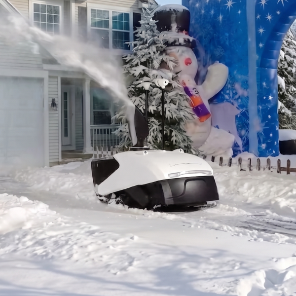 White and black robotic snow blower clears driveway, spraying snow; inflatable snowman and blue arch in background, sunny winter day.