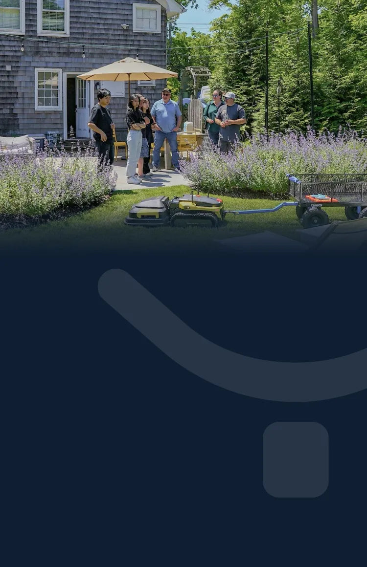 Group gathers in sunny backyard with purple flowers, yellow umbrella, and robotic lawn mowers on grass.