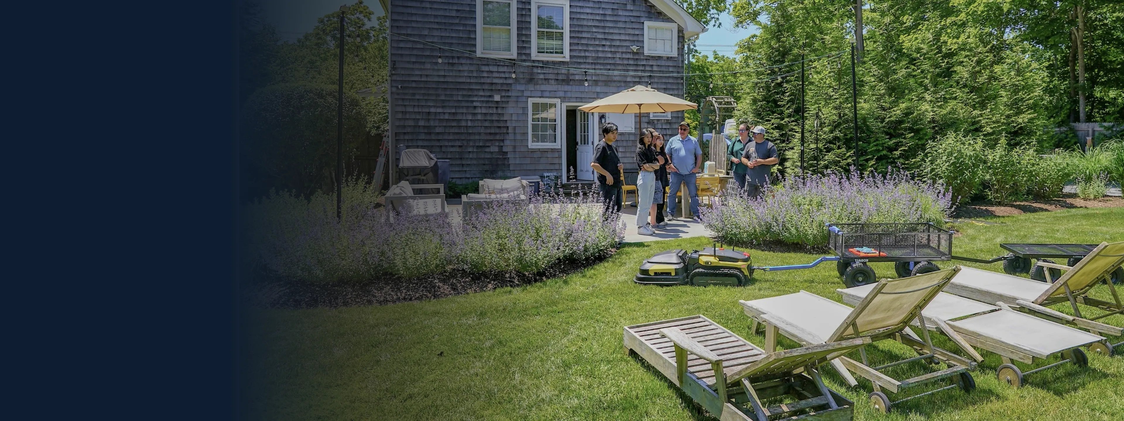 Group gathers on sunny patio beside gray shingled house, with lawn chairs, purple flowers, and robotic mowers on green grass.