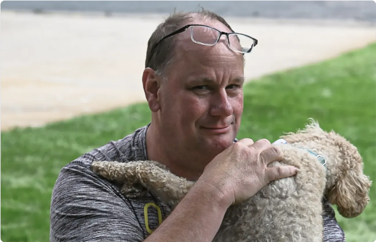 Man with glasses on head smiles while holding fluffy light-brown dog in grassy outdoor setting.