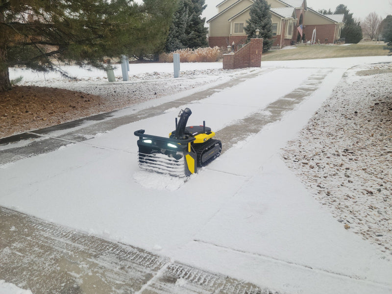 Yellow and black robotic snow plow clearing driveway, snow-covered yard, house in background.