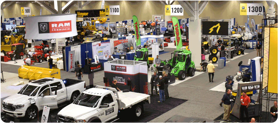 Wide indoor trade show floor with white RAM trucks, green tractors, and branded booths under bright lighting.