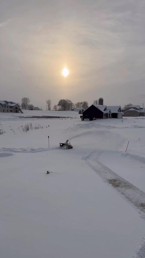 Snow-covered farmyard with dark barn, silo, and house under bright sun; snowblower clears path in foreground.