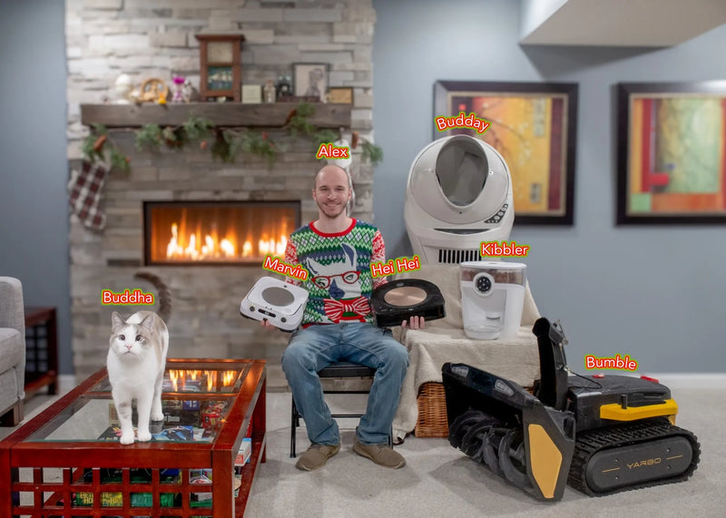 Man in festive sweater sits with robot vacuums and cat in cozy living room with fireplace.