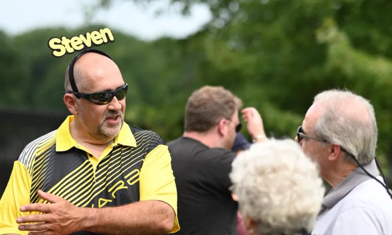 Man in yellow and black striped polo shirt labeled "Steven" wearing sunglasses, speaking to others outdoors.