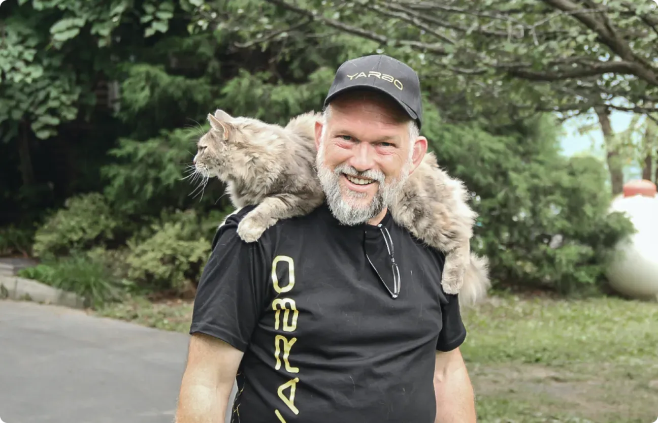 Smiling bearded man in black cap and shirt with fluffy gray cat on shoulders, outdoors in greenery.