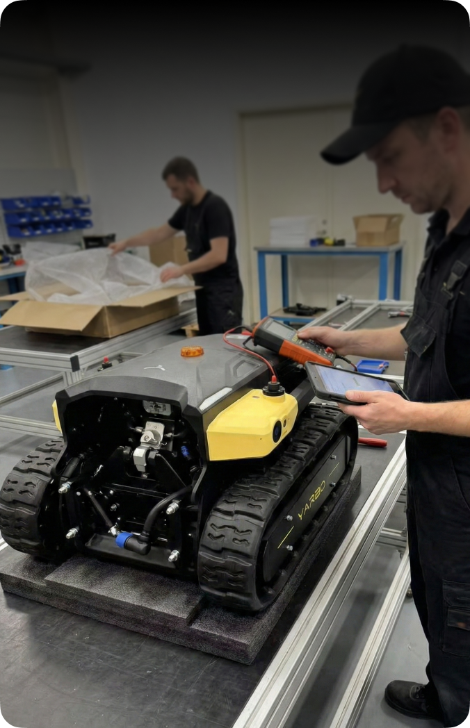 Black and yellow tracked robot on workbench, technician using tablet and multimeter, another unpacking box in background.