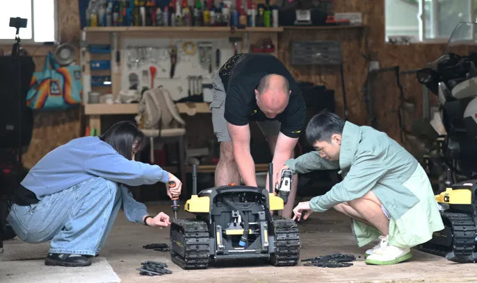 Three people assemble a black and yellow tracked robot in a workshop, surrounded by tools and shelves.