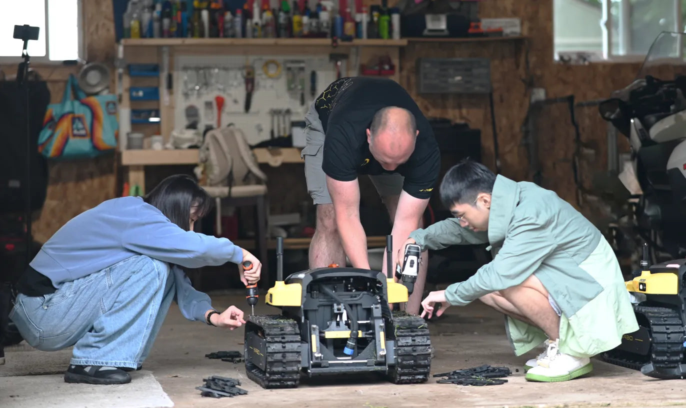 Three people crouching in a workshop, repairing a black and yellow tracked robot with tools, surrounded by scattered parts and equipment.