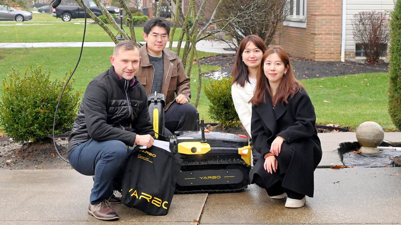 Four people pose with a black and yellow YARBO robotic lawn mower on a sidewalk, surrounded by greenery and a brick building.