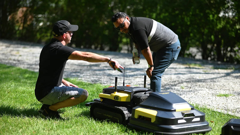 Two men inspect black and yellow robotic lawn mowers on grass, one crouching, one bending, under bright sunlight.