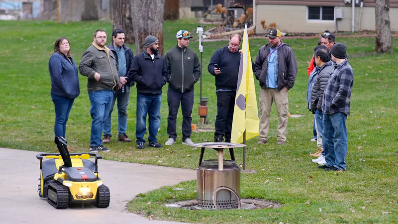 Yellow tracked robot on sidewalk near group of people observing in grassy yard with metal fire pit and yellow flag.