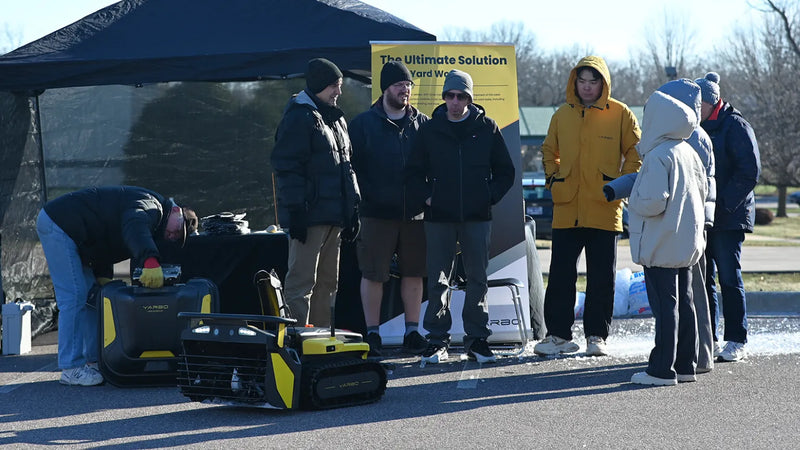 People gather outdoors near a black and yellow YARBO robotic lawn mower and branded tent under bright sunlight.