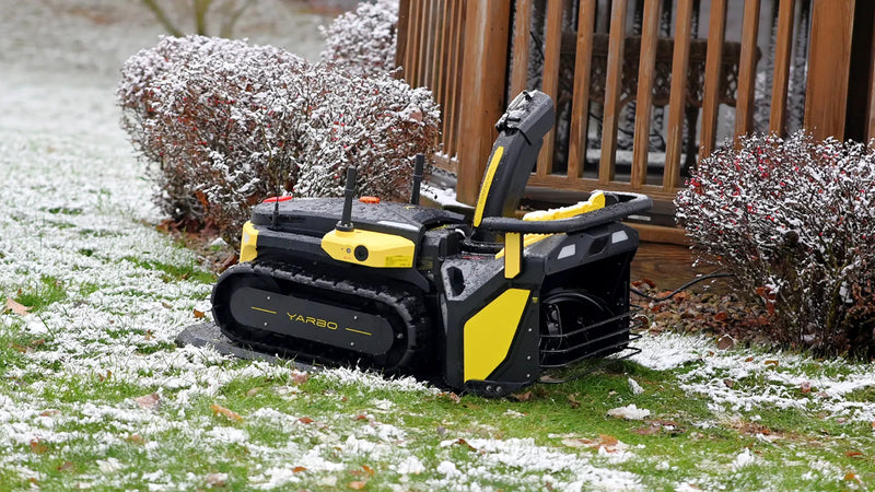 Black and yellow YARBO robotic lawn mower on snowy grass near wooden fence and snow-dusted bushes.