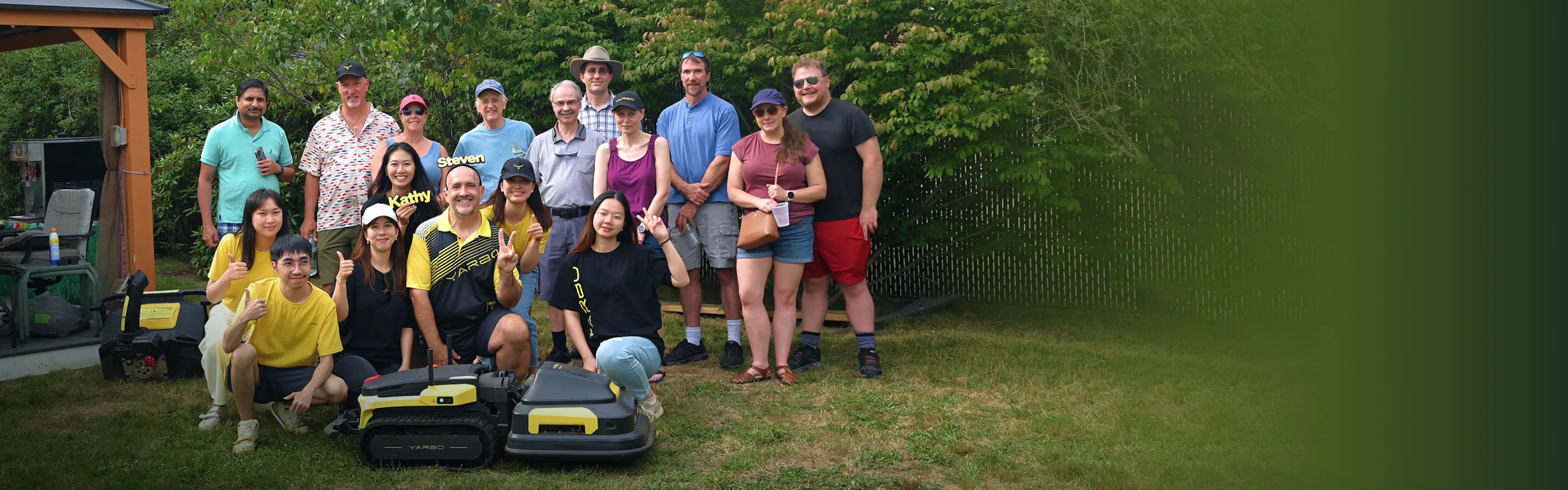 Group of people posing outdoors with two YARBO robotic lawn mowers on grass, smiling and gesturing.