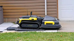 Black and yellow robotic lawn mower with tracks, parked on concrete patio near brown siding and green grass.