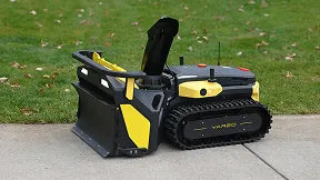 Yellow and black YARD BOT robotic lawn mower on concrete, grass background, tracks visible, compact design.